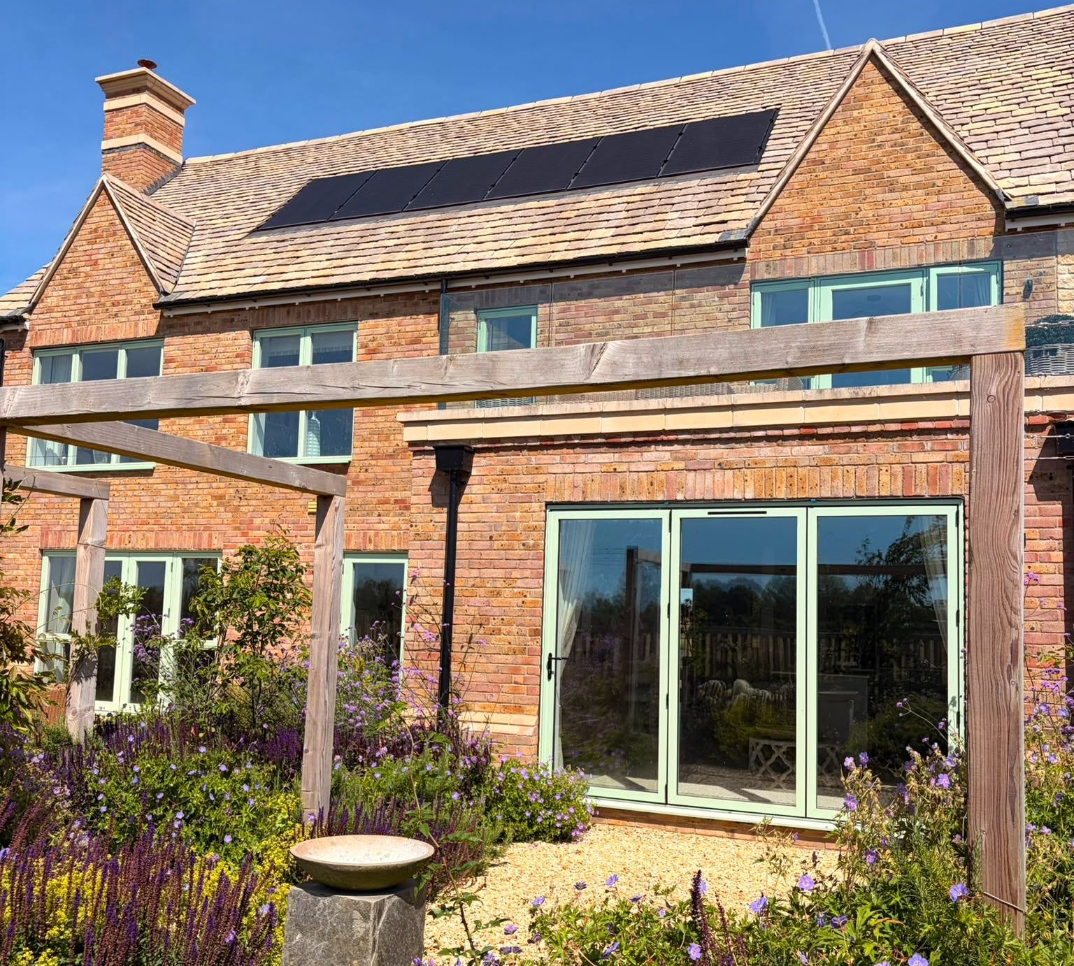 tunning brick house featuring modern solar panels, sage green windows, a beautiful garden with purple flowers, and a charming pergola. Sustainable living in style.
